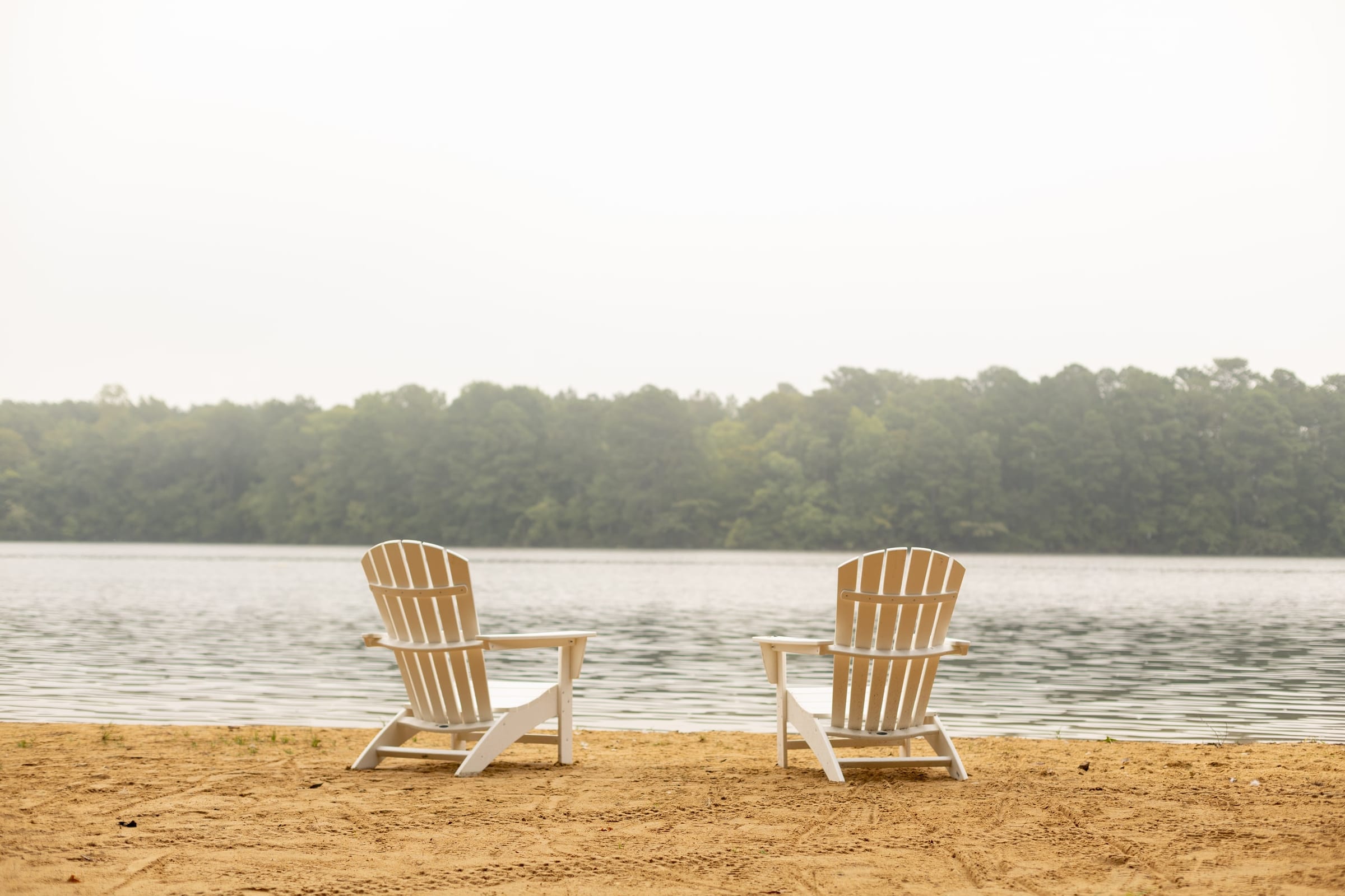 Two white Adirondack chairs on the sand beach facing the hazy lake