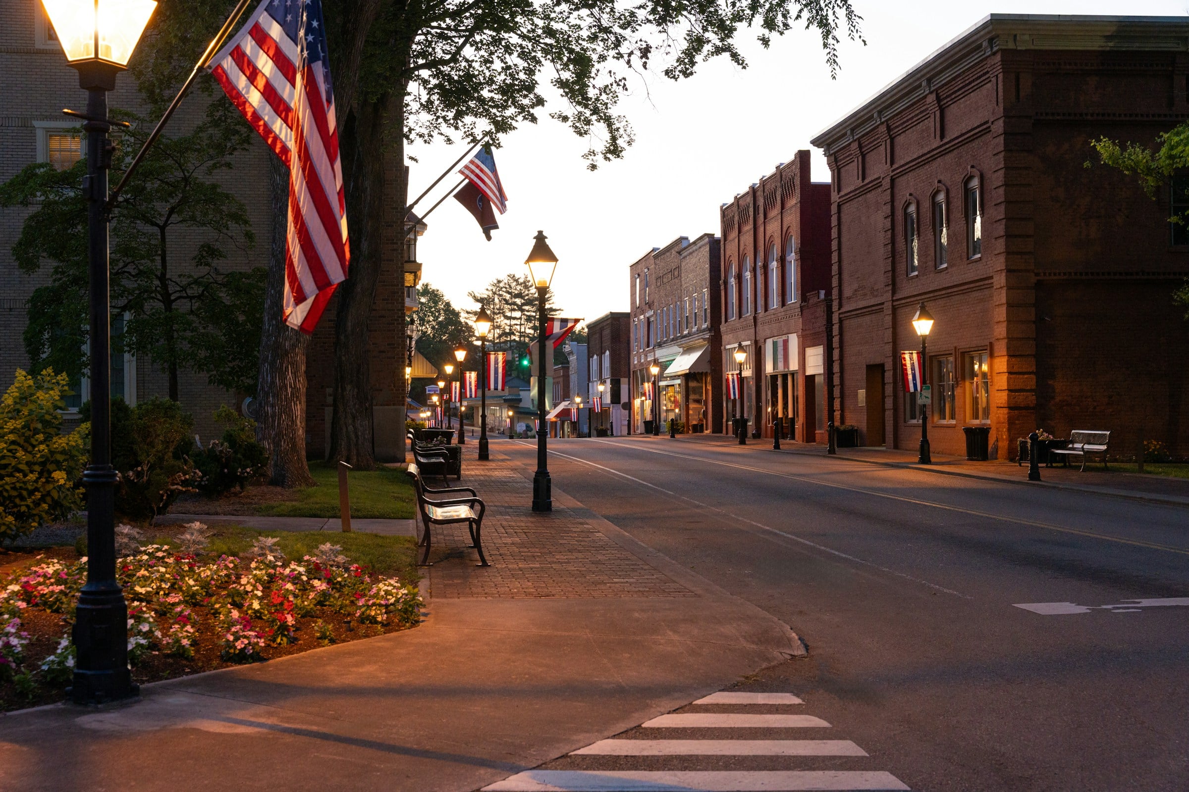 Small-town main street at dusk: brick storefronts, glowing lampposts, American flags, benches