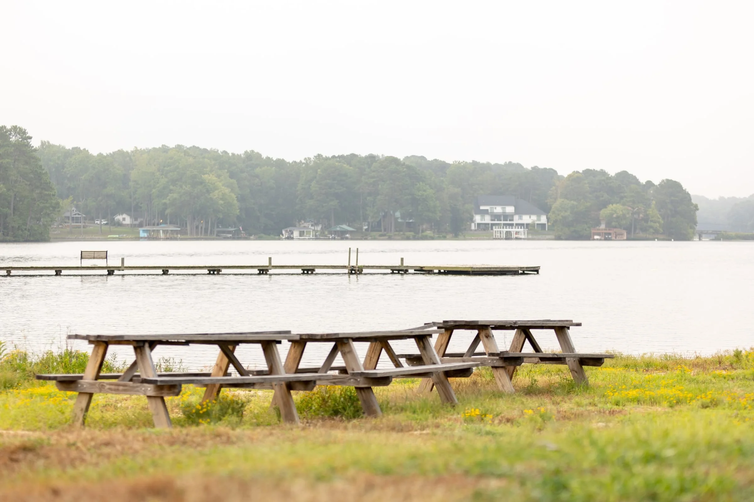 Wooden picnic tables on the waterfront lawn with docks and the hazy far shore behind